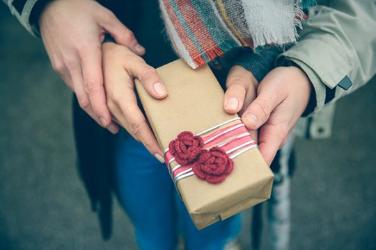 Closeup of woman and man hands showing a gift box with red handmade flowers otdoors in a cold autumn day. Love and couple relationships concept.